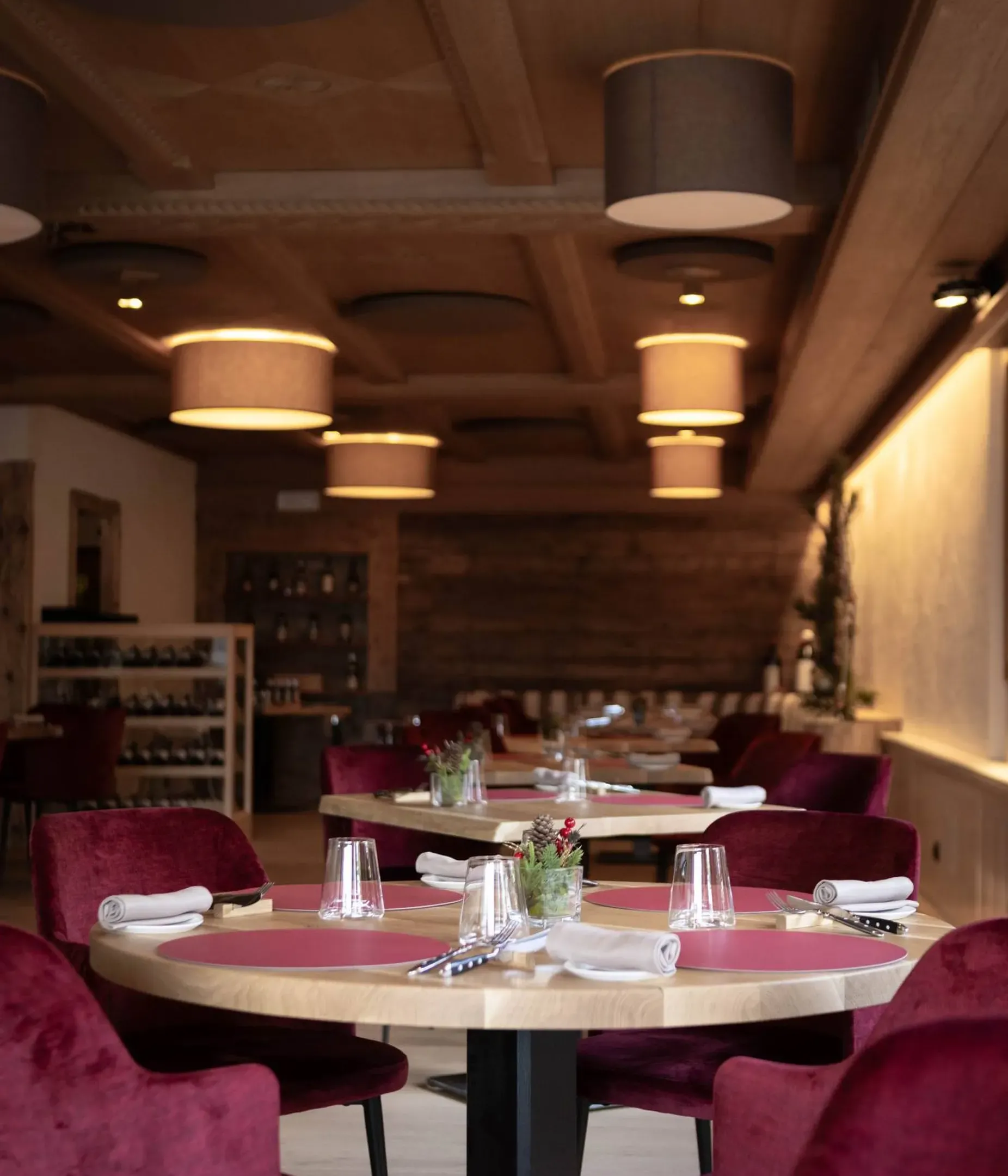 Interno di ristorante elegante con tavoli rotondi di legno, sedie velluto rosso, tovaglioli arrotolati e vasi di piante, luci pendenti calde.